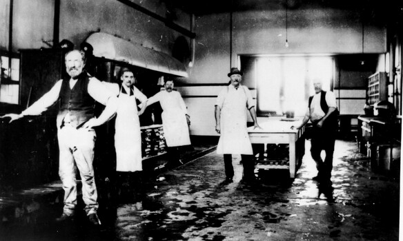 A group of five men standing in a kitchen. Three men are wearing aprons indicating they may be the cooks.