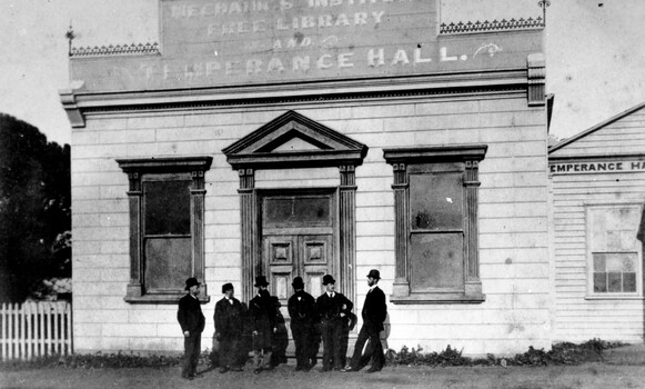 A group of men in suits and hats are standing outside the front entrance of the Mechanics Institute and Temperance Hall, Cheltenham