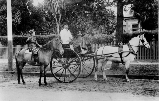 A man wearing a white coat and a traditional butcher's apron is sitting in the buggy of a horse drawn wagon. A young boy is sitting on a horse beside him.