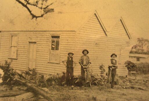 Sepia image of three men standing in front of a white weatherboard building, two men leaning on pitchforks