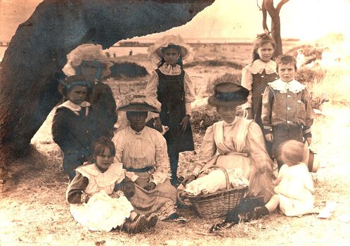 Sepia image of a family with two women and children sitting on grass under a tree with a picnic basket