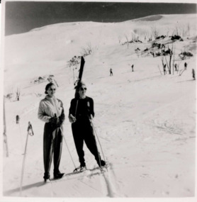 Two women in ski clothing and holding skis in foreground with white snowy hill and skiers in background. Small amount of sky in background. 