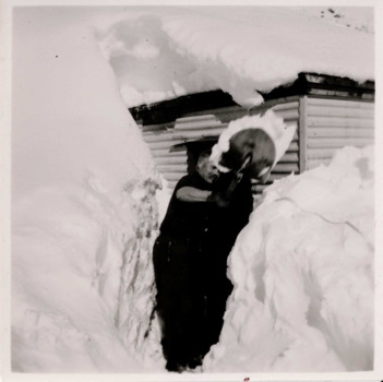 Man shovelling snow in front of himself. High snow banks either side of man and and on the building. 