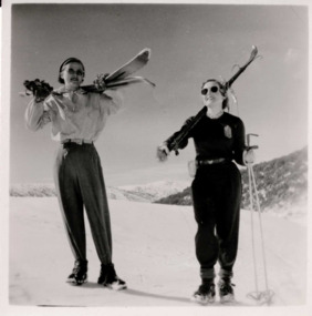 Two women in snow smiling, wearing ski attire and holding skis over their shoulders.
