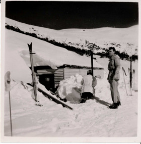 Two people in front of a hut. One person is sunken in the snow and the person is higher up on snow.  
