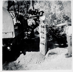 A man wearing a hat behind and operating machinery. A child standing to the right. Trees in the background. 
