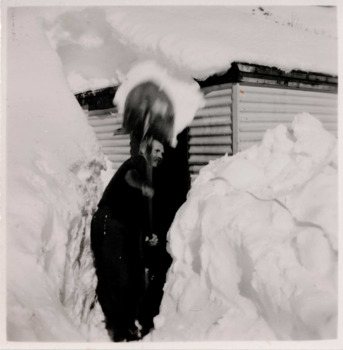 Man shovelling snow at centre with high levels of snow either side of him and a snow-covered hut in background. 