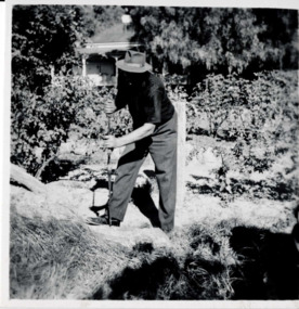 Man holding a long tool above ground. He is wearing a hat shadowing his face. Trees and a house are in the background. 
