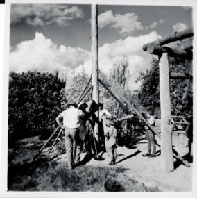 3 men and 2 children with a tall wooden structure over a hole in the ground. A wooden building structure is to the right.