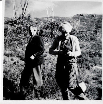 Photograph of two people standing outside: one person facing left and walking while the other is to their right facing forward and looking down