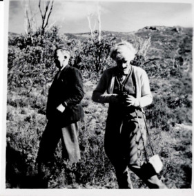 Photograph of two people standing outside: one person facing left and walking while the other is to their right facing forward and looking down