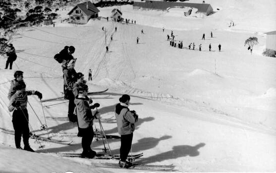 A group of skiers at the top of a slope looking down towards other skiers and the lodge below.