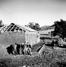 Four men standing at the front of a building under construction. Small tractor to the right of the building.