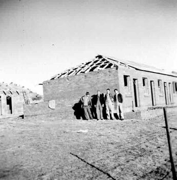 Four men in front a building under construction. Walls are standing but roof is in progress,