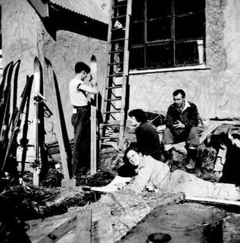 Four people outside the hut. One is standing and working on a ski, the remainder are seated.