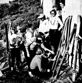 People at the front of the hut, two women in foreground are examining skis leaning against the wall.