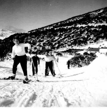 A group of skiers ascending the slope with taller tree lined slope in background. Building in background to the right.