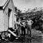 Three people beside building. Woman is pointing to the right. Bucket and container beside step.
