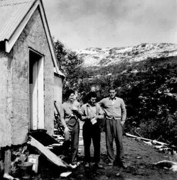 A Woman and two men beside building. Bucket and container beside step.
