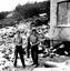 Three people standing beside building. Small machinery in background.