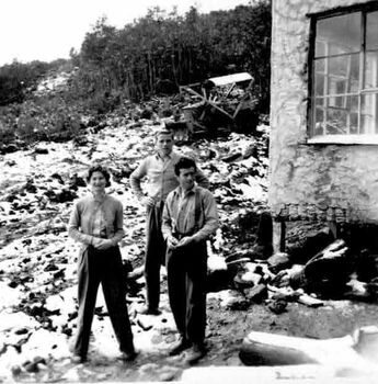 Three people standing beside building. Small machinery in background.