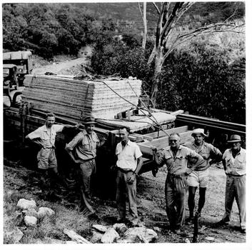Six men with timber loaded truck. Other vehicle in background on left.