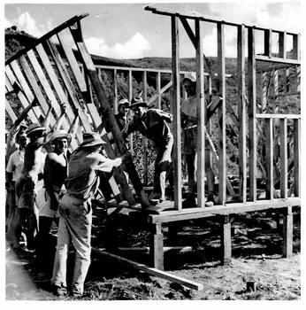 Another section of frame being lifted into place. 3 men inside the building and others raising the frame from the ground.