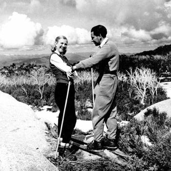 An unnamed couple on skis with mountain views and clouds in the background. Woman is looking towards the camera.