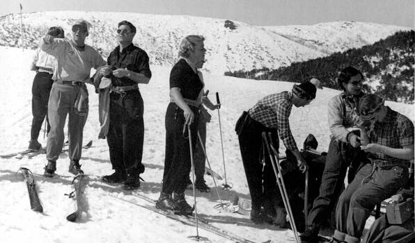 A group of skiers on the snow. One man on left pointing down the mountain. Large amounts of snow on the ground.