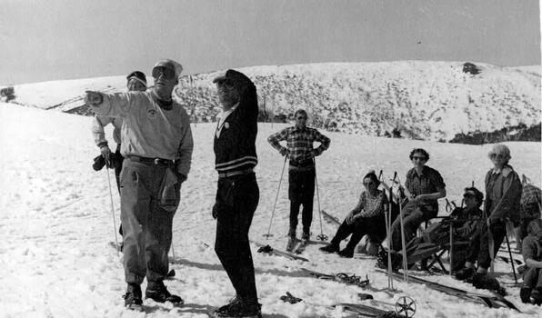 Group of skiers seated to the right, two men in foreground looking down the slope with man furthest left pointing into the distance.