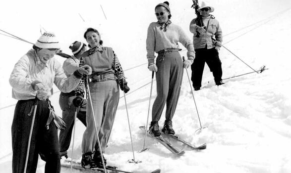 Five skiers on the slopes, two women in foreground are laughing whilst others watch on.