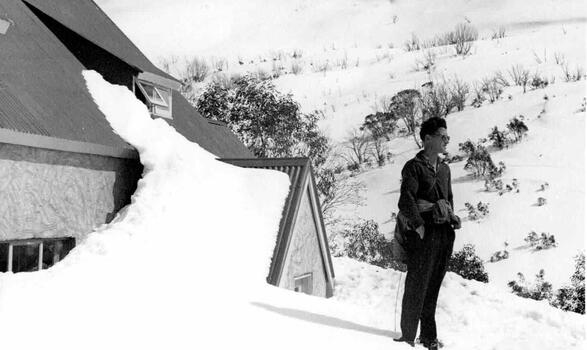 A heavily snow-covered Bogong lodge with a person standing to the right and looking into the distance.