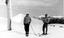 Two men on skis standing beside a pole. A thick covering of snow around them.