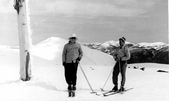 Two men on skis standing beside a pole. A thick covering of snow around them.