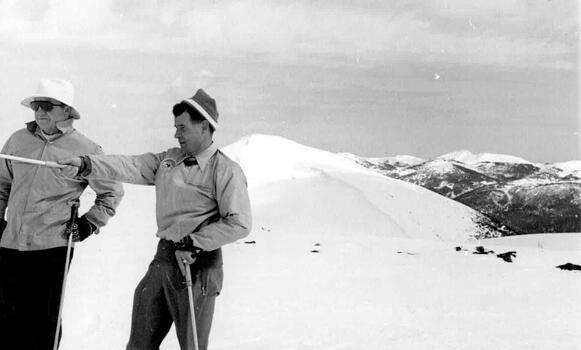 Two men on left foreground looking and pointing to the left. Snow covered mountains in background