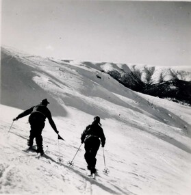 Two skiers heading away from the camera towards snow covered hills.