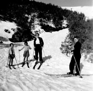 Two adults and two skiing on the road near the lodge. Children are wearing jodhpurs.