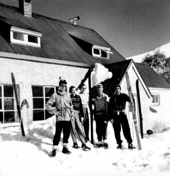 Five people with skis in front of lodge in heavy snow. One set of skis against wall
