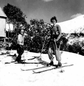 A woman and  child on skis. Corner of building visible on the left and snow covered slope on the right.