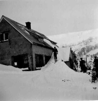 A man standing on ramp formed by snow from verandah roof and another man on the right.