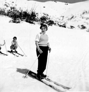 A woman standing on skis, another woman squatting on skis behind her with a third person cut off at left edge of image