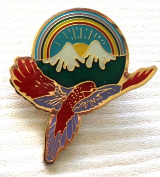 A badge depicting the sun shining behind 2 mountains. The lower edge features a bird with wings extended and a rainbow circles the centre,
