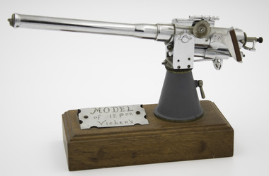 Rectangle wooden base with model gun mounted on one end with silver engraved label screwed on top of base next to base of gun model.