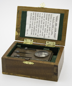 Rectangular honey coloured wooden box with hinged lid, open, shows printed paper history about the item in lid and metal torch in box.