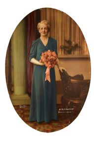 Oval shaped studio portrait depicting a standing woman in a green evening dress wearing white gloves and pearls holding a bouquet of pink flowers. Woman has her left hand on the top back of a chair. 	