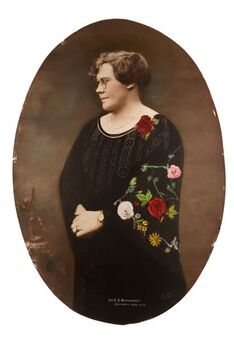 Three quarter oval shaped studio portrait depicting a standing woman in profile view wearing a dark evening dress and pearls. 