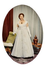 Oval shaped studio portrait depicting a standing woman in a white evening dress wearing white gloves and the gold Mayoress medallion. Woman has her right hand on the top back of a chair. 