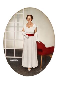 Oval shaped studio portrait depicting a standing woman in a white evening dress with a red sash wearing white gloves and the Mayoress medallion.  The woman has her left hand on the top back of a chair. 