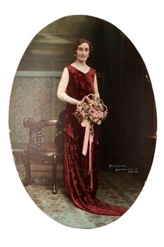 Oval shaped full length studio portrait depicting a standing woman in a red evening dress wearing white gloves and  holding a bouquet of pink flowers. 