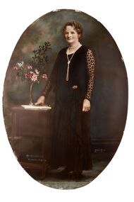 Oval shaped full length studio portrait depicting a standing woman in a dark evening dress with lace sleeves wearing pearls. The woman has her right hand on the top of a small table next to a small vase of flowers.